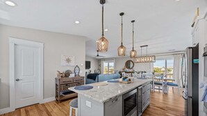 A modern kitchen with a central island, wooden flooring, pendant lighting, and a view to the outside. - 3943 - Let it Beach - Walk to Duck Shops + Beach | Pool & Hot Tub (Duck)