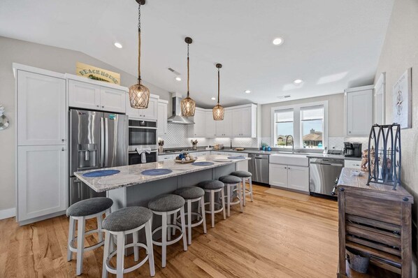 A modern kitchen with white cabinets, stainless steel appliances, a central island with bar stools, pendant lighting, and a view to the outside.