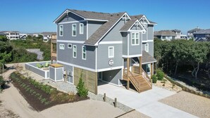 A two-story house with a covered porch and a small deck, surrounded by a landscaped garden and a sandy driveway. - 3943 - Let it Beach - Walk to Duck Shops + Beach | Pool & Hot Tub (Duck)