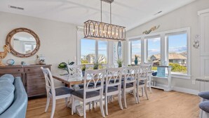A dining area with a wooden table, white chairs, a chandelier, and large windows offering a view of the neighborhood. - 3943 - Let it Beach - Walk to Duck Shops + Beach | Pool & Hot Tub (Duck)