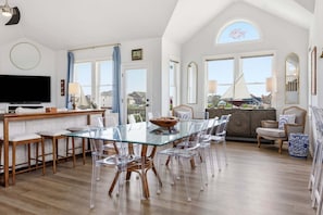 A spacious dining area with a glass table and clear chairs, a wooden bar, and a large TV.