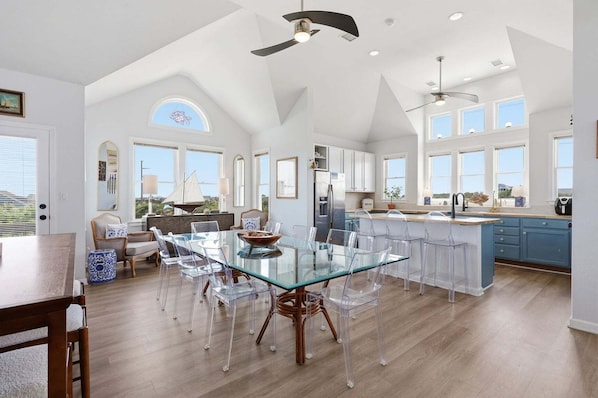 A spacious kitchen with a glass dining table, clear chairs, and a high ceiling with a fan.