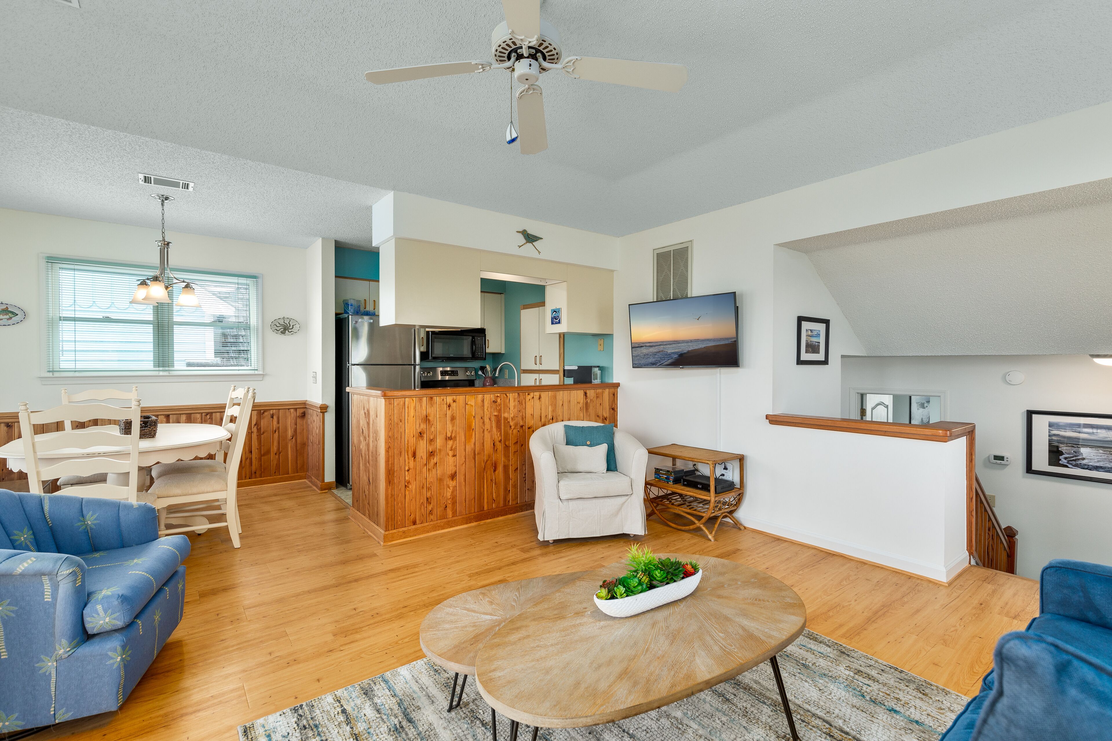 A spacious living area with a wooden dining table, two blue sofas, a coffee table with a plant, a kitchen area with a microwave, and a TV mounted on the wall.