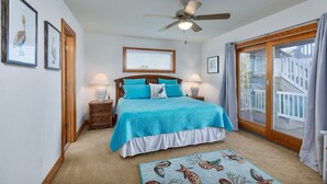A bedroom with a bed, two bedside tables, a ceiling fan, a window with blinds, and a view of the outside deck. - 5788- Birds Eye View - Beach Getaway in Nags Head | Private Pool (Nags Head)