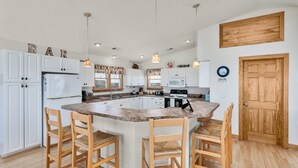 A modern kitchen with white cabinets, a central island with bar stools, pendant lighting, and a wooden door. - 5788- Birds Eye View - Beach Getaway in Nags Head | Private Pool (Nags Head)
