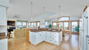 A spacious kitchen with white cabinets, a wooden countertop, and a large island. There is a dining area with a table and chairs, and a living room with a sofa and a TV. - 5788- Birds Eye View - Beach Getaway in Nags Head | Private Pool (Nags Head)