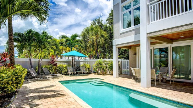 A swimming pool with lounge chairs and umbrellas, surrounded by palm trees and a white building with large windows. - NO Bunks-King beds, Garage Parking, Private Beach Access-Less Crowded (Siesta Key)