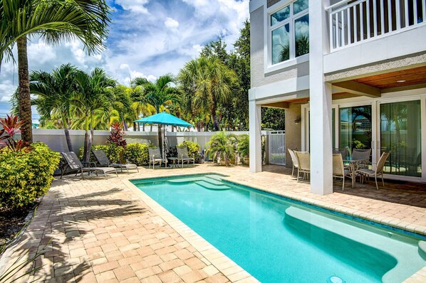 A swimming pool with lounge chairs and umbrellas, surrounded by palm trees and a white building with large windows.