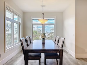 Un espace repas comprenant une table et des chaises en bois, un luminaire suspendu et de grandes fenêtres donnant sur l’extérieur.