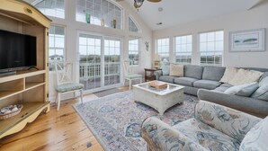 A spacious living room with a large flat-screen TV, a ceiling fan, and a large window overlooking a deck. - 4763- Smells Like Sea Spirit, Kitty Hawk Beach House | Pool & Game Room (Kitty Hawk)