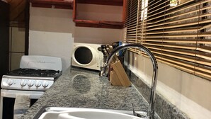 A compact kitchen with a gas stove, oven, and a built-in microwave. There is a sink with a modern faucet, and wooden shelves above the counter. - Villa by the Sea (Playa Grande)