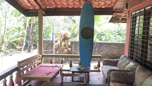 A covered outdoor seating area with a table, chairs, a surfboard, and a potted plant. - Villa by the Sea (Playa Grande)