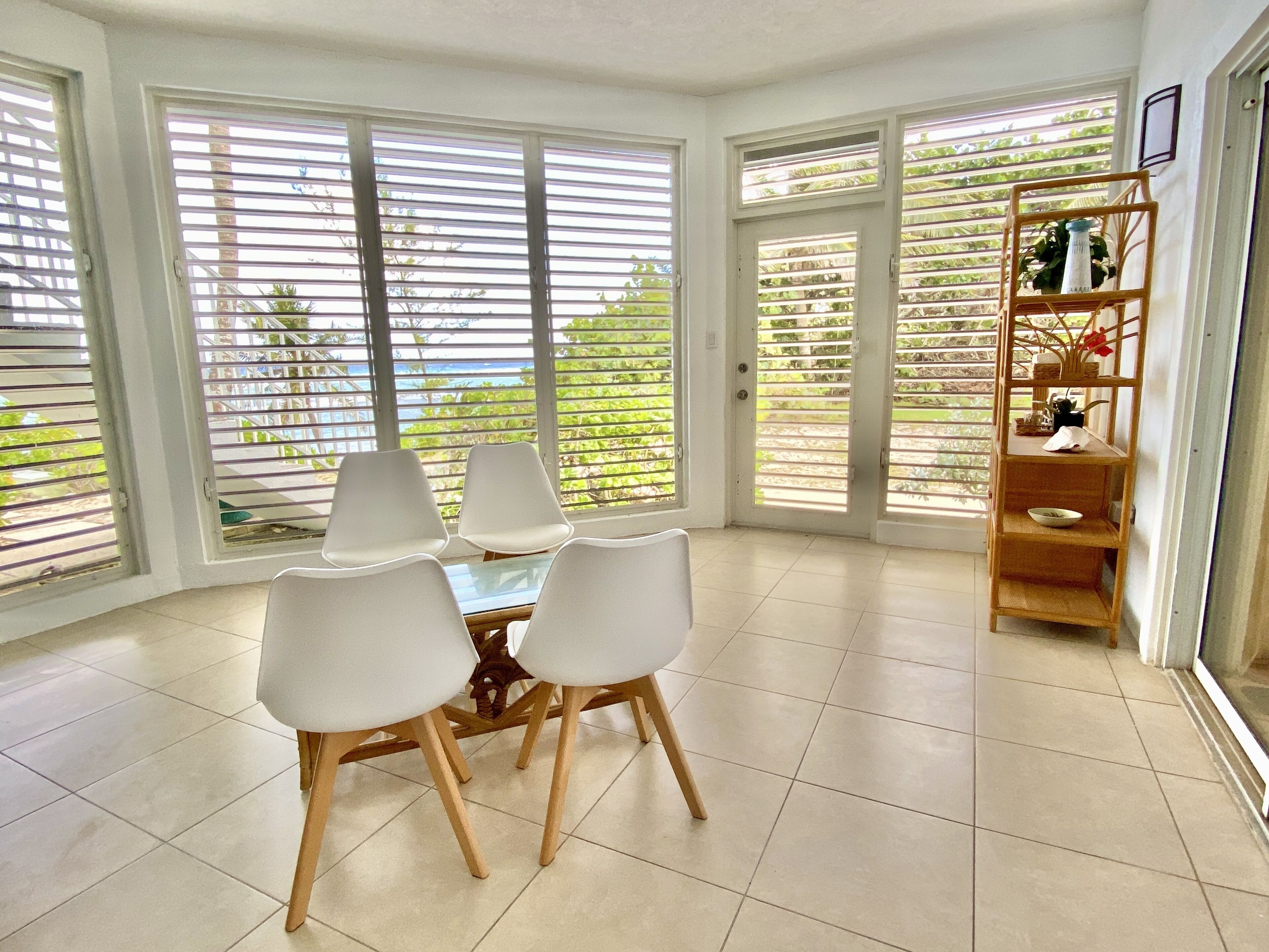 A dining area with a table and chairs, large windows with blinds, and a wooden shelf with decorative items.