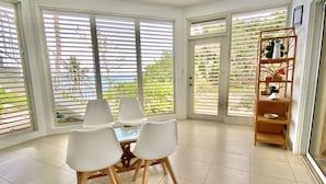 Un coin repas avec une table et des chaises, de grandes fenêtres équipées de stores et une étagère en bois agrémentée d’objets décoratifs.