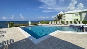 A swimming pool with a view of the ocean, surrounded by lounge chairs and a building with a green roof.