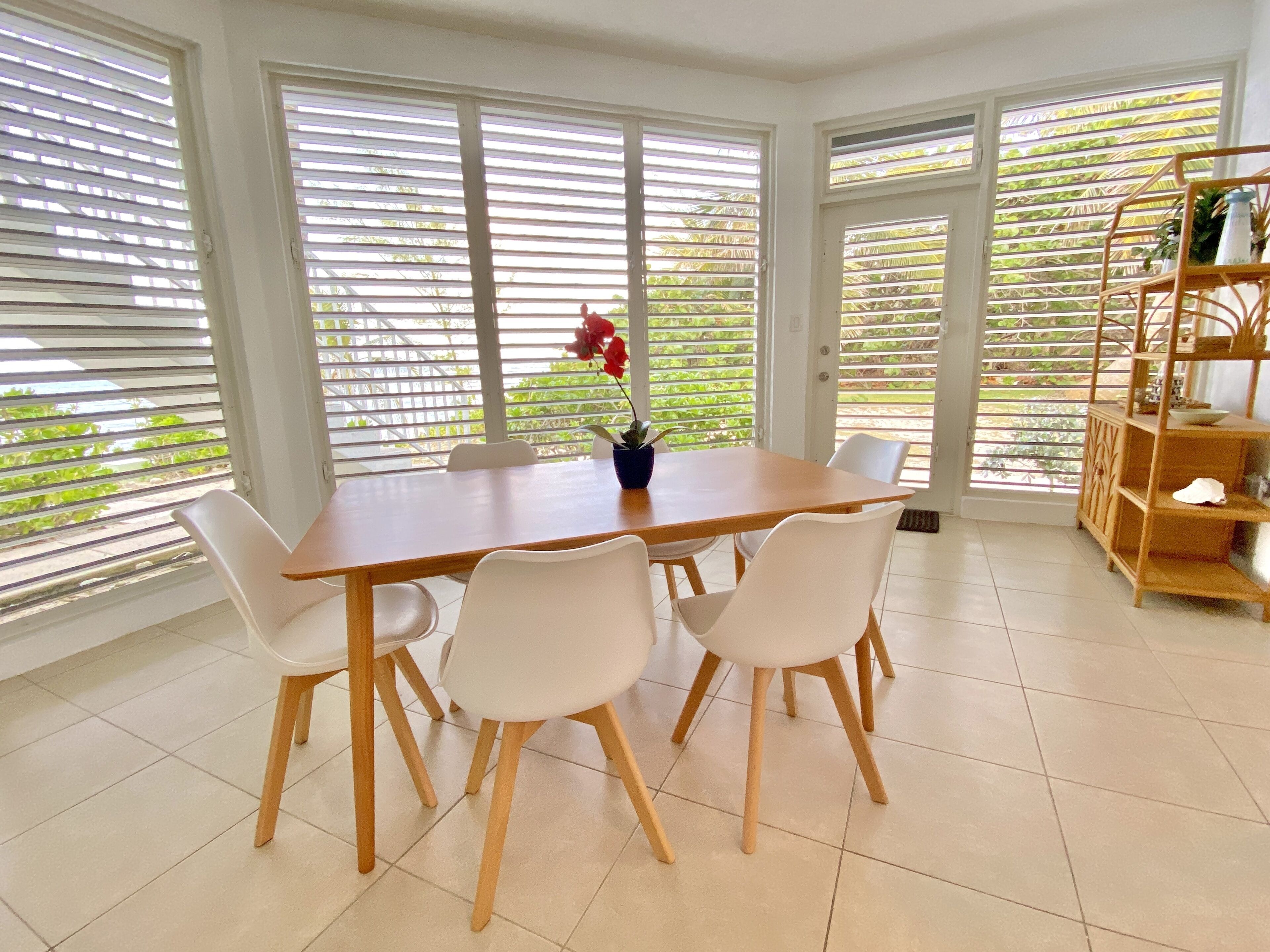 Een eetgedeelte met een houten tafel en witte stoelen, grote ramen met jaloezieën en een houten wandplank met planten.
