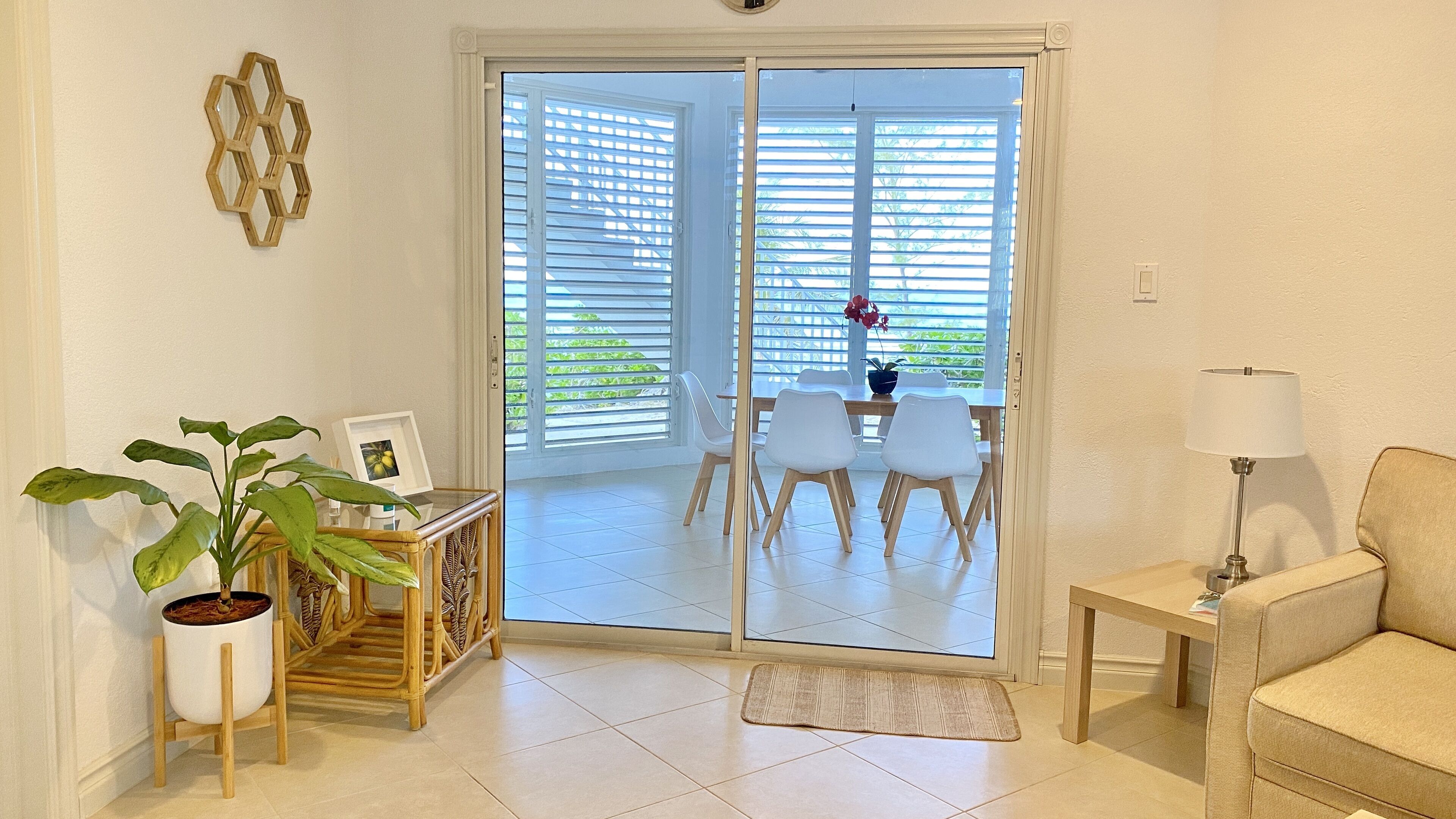 Un salón con un sofá, una mesa con sillas, una planta en maceta, una lámpara y un ventanal corredero que da a un balcón con mesa y sillas.