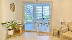 Un salon comprenant un canapé, une table avec des chaises, une plante en pot, une lampe et une porte-fenêtre coulissante donnant sur un balcon équipé d’une table et de chaises.