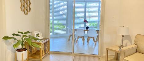 Un salón con un sofá, una mesa con sillas, una planta en maceta, una lámpara y un ventanal corredero que da a un balcón con mesa y sillas.