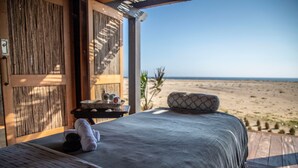 Un dormitorio con vista al mar, una puerta de madera, una mesita con artículos de aseo y una cama con manta y almohadas texturizadas.
