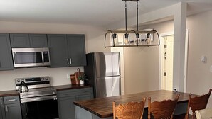 A modern kitchen with grey cabinets, stainless steel appliances, a wooden dining table, and a trash can. - Newly remodeled Riverfront 3 bedroom house on the Great Chazy River with dockage (Champlain)