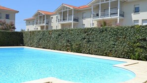 A swimming pool with a tiled edge, surrounded by a hedge and adjacent to a building with balconies. - Beautiful apartment for 4 people (BISCARROSSE)