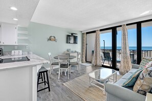 A modern kitchen with a marble countertop, a dining area with a round table and chairs, a sofa with patterned cushions, and a view of the ocean through large windows.
