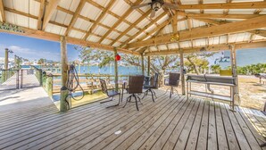 Una terraza de madera con sillas y mesas de metal, un ventilador de techo y vista al agua.