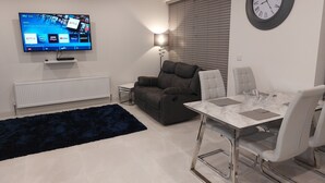 A modern living room with a flat-screen TV, a sofa, a dining table with chairs, and a wall clock.
