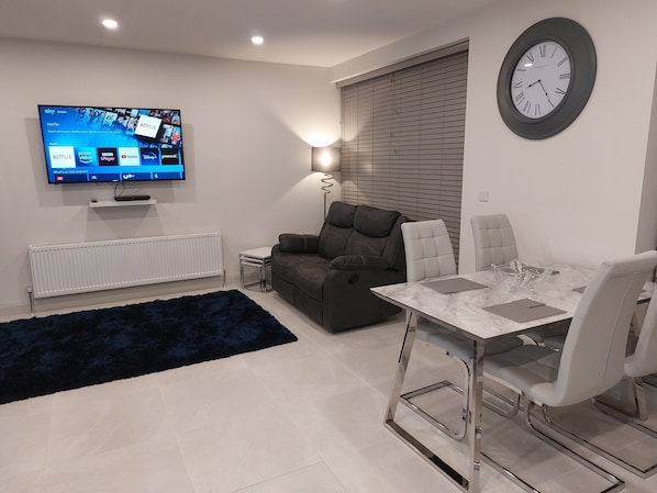 A modern living room with a flat-screen TV, a sofa, a dining table with chairs, and a wall clock.