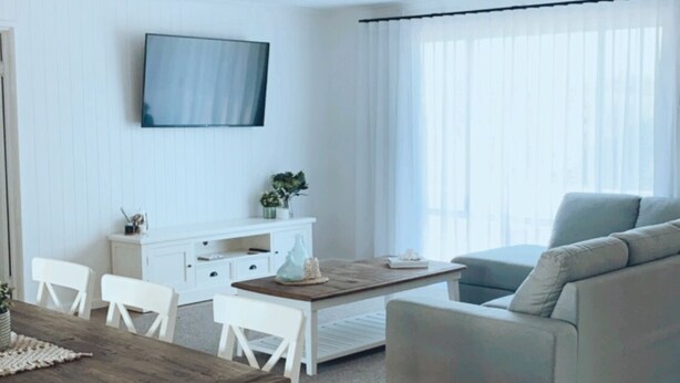 A modern living room with a wooden dining table, white chairs, a grey sofa, and a flat-screen TV mounted on the wall.