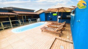 Area piscina sul tetto con lettini in legno e ombrelloni, una piscina e vista su edifici dalle pareti blu e tetti in tegole.