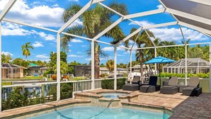 A pool area with a clear view of the surrounding landscape, including palm trees and a distant building. - Luxury home w/ 2 suites, LARGE pool, spa, lanai. Quick gulf access! 6 Bikes! EV! (Cape Coral)