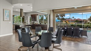 A modern kitchen with dark cabinets, a dining area with a glass table and chairs, and a patio with outdoor seating and a view of a building with a red roof. - Luxury home w/ 2 suites, LARGE pool, spa, lanai. Quick gulf access! 6 Bikes! EV! (Cape Coral)