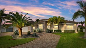 A modern house with a paved driveway, palm trees, and a well-maintained lawn. - Luxury home w/ 2 suites, LARGE pool, spa, lanai. Quick gulf access! 6 Bikes! EV! (Cape Coral)