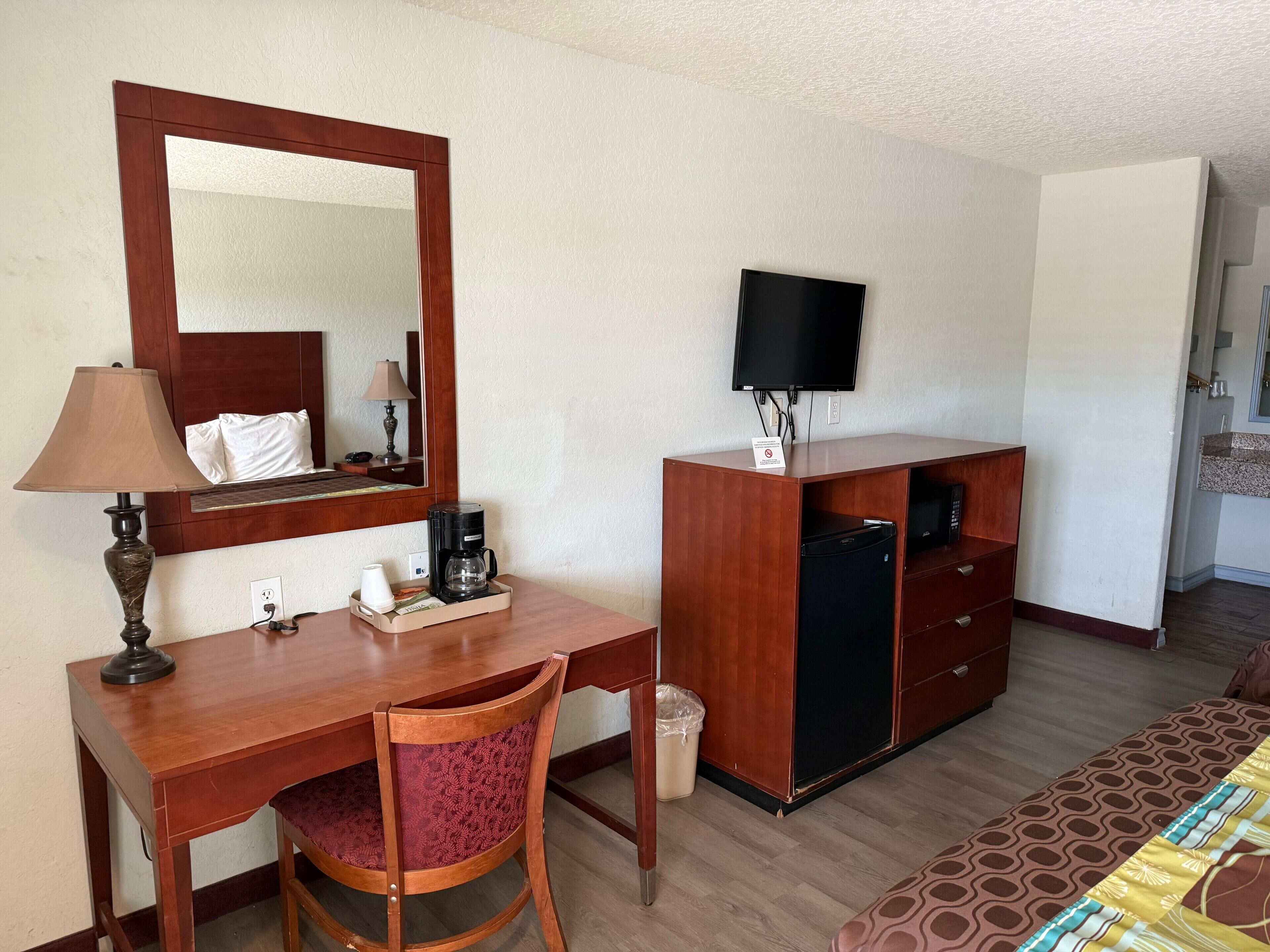 A hotel room with a wooden desk, a chair, a bed with a patterned bedspread, a television, a coffee maker, and a small trash bin.