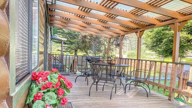 A covered outdoor patio with a table and chairs, a bench with flowers, and a wooden railing. - Meadowview Clean & Comfortable Home by River Side (Klamath)