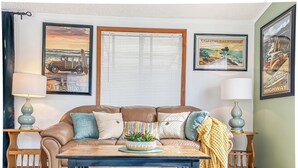 A living room with a brown leather sofa, a blue coffee table, and a small crab figurine. There are two lamps on wooden side tables, a window with blinds, and framed pictures on the walls. - Meadowview Clean & Comfortable Home by River Side (Klamath)