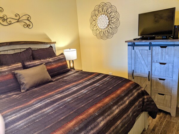 A bed with a striped blanket, a decorative wall piece, a television on a wooden cabinet, and a lamp on a bedside table.