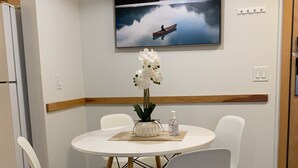 A dining area with a round table, white chairs, a vase of white orchids, and a painting of a lake with a boat. - Gateway to Rocky Mountains -1 Bdr Suite/Kitchen/AC/UG Pk/Gym (Canmore)