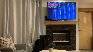 A living room with a television, a fireplace, a sofa, and a small table. - Gateway to Rocky Mountains -1 Bdr Suite/Kitchen/AC/UG Pk/Gym (Canmore)
