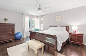 A bedroom with a wooden bed, a nightstand with a lamp, a dresser, a blue exercise ball, a fluffy stool, and a ceiling fan.