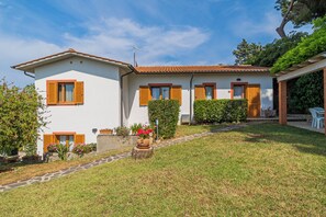 Une maison à deux étages, avec un toit en tuiles, des murs blancs et des volets en bois. Un chemin pavé mène à l’entrée, et un petit jardin agrémenté de plantes en pot et de fleurs.