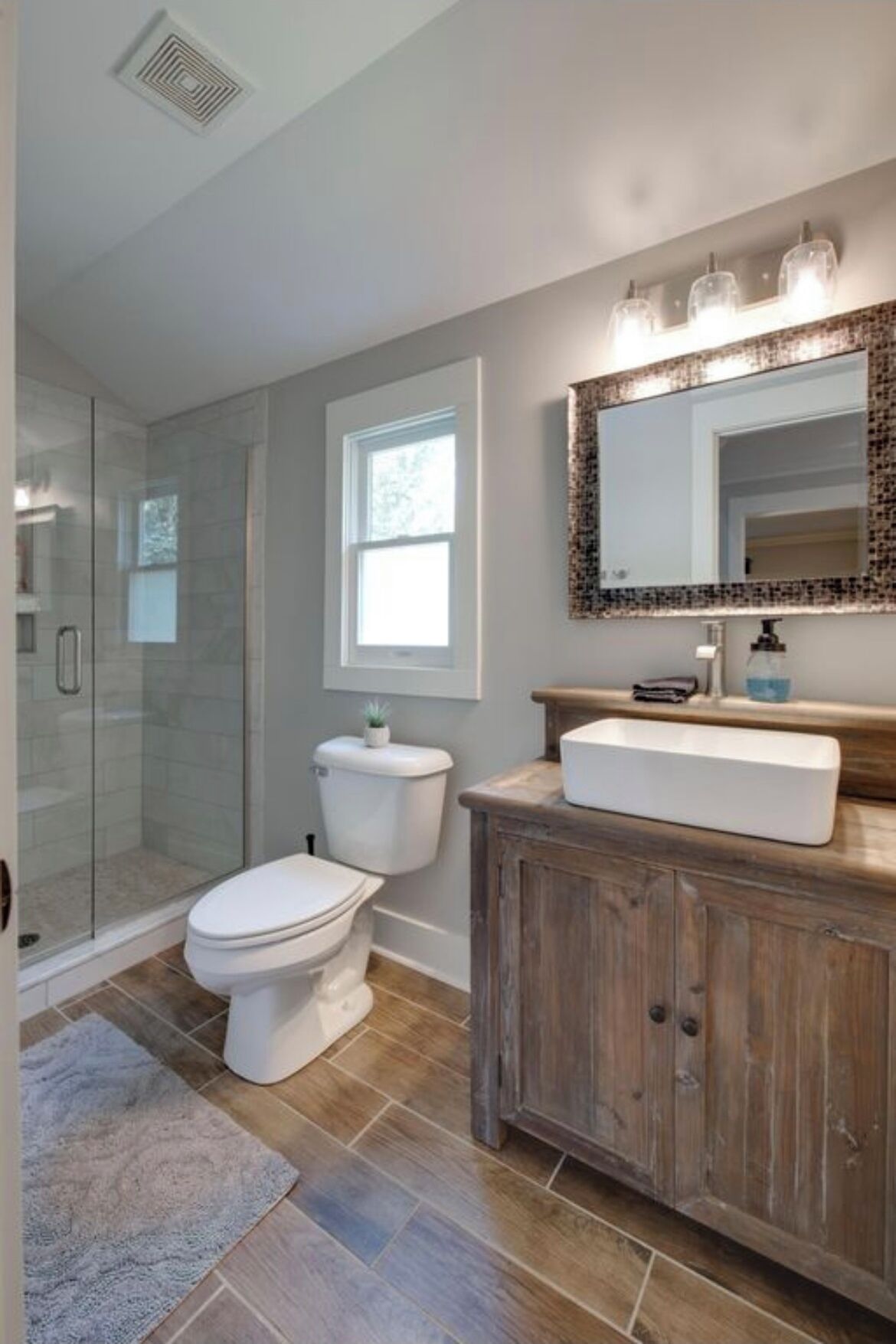 Une salle de bain dotée d’une douche à parois de verre, de toilettes blanches, d’un meuble-lavabo en bois et d’un miroir avec un cadre décoratif.