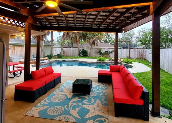 Une terrasse extérieure couverte avec une piscine, des sièges rembourrés de couleur rouge et un tapis à motifs.