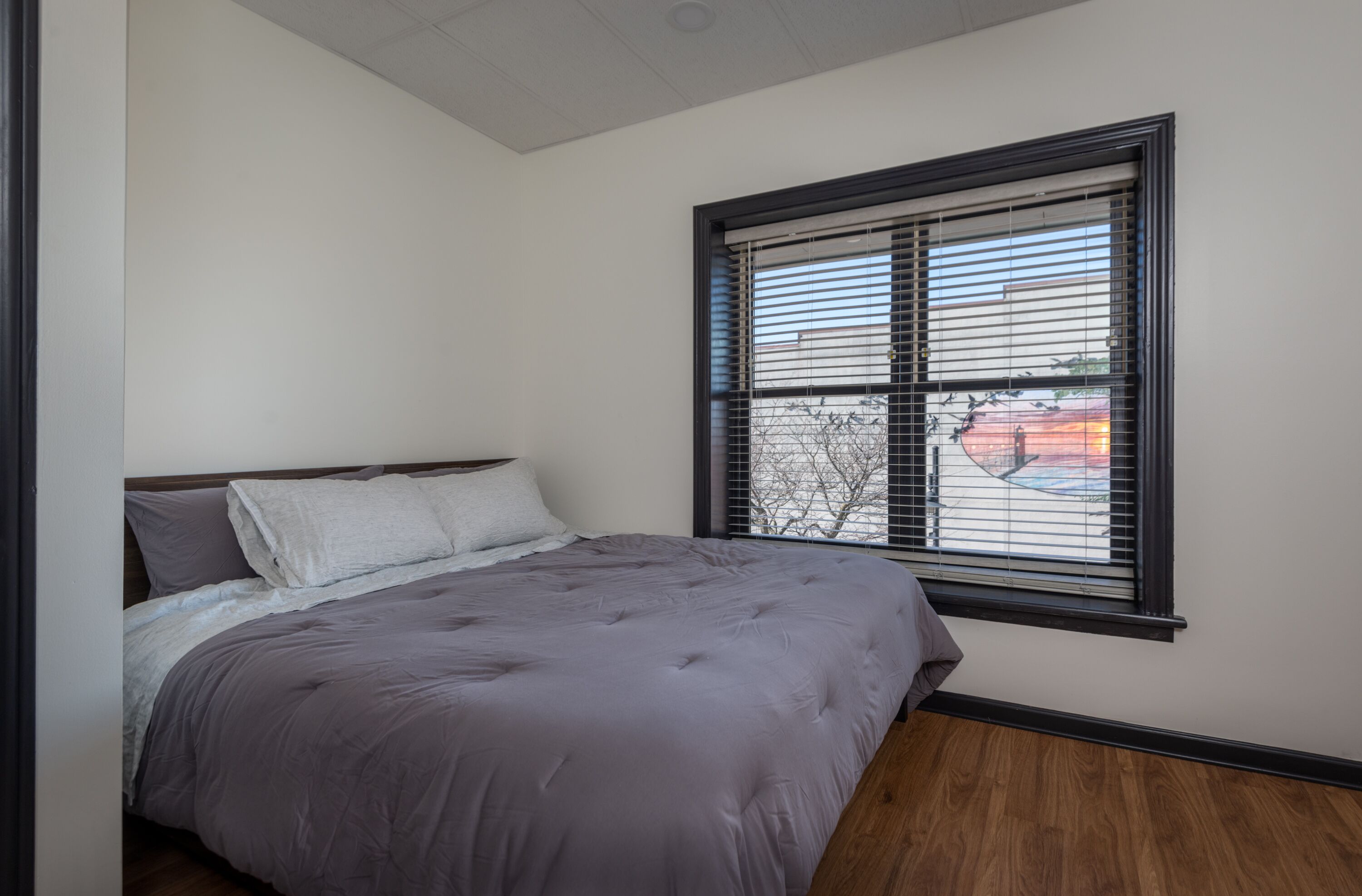 Ein Schlafzimmer mit Bett, einem Fenster mit Jalousien und Blick ins Freie.