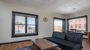 A modern living room with a sofa, a wooden coffee table, and a rug. There are two windows with blinds, a clock on the wall, and a view of buildings outside.