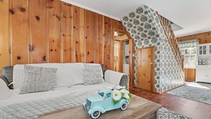 Een gezellige woonkamer met een houten wand, een bank met gestreepte kussens, een klein tafeltje met een speelgoedauto en bloemen, en een trap met behangpapier met een patroon.