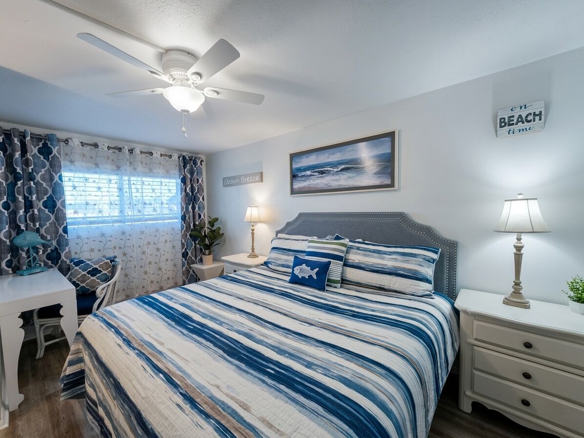 Une chambre à coucher comprenant un lit, des tables de chevet, un bureau, un ventilateur de plafond et un panneau décoratif mural sur le thème de la plage.