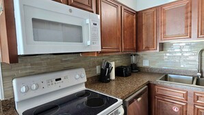 A kitchen with wooden cabinets, a white microwave, a stove, and a sink. - Beachfront Condo w/ FREE Beach Service, 2nd Floor, Pools, Saunas, Tootsie's (Panama City Beach)